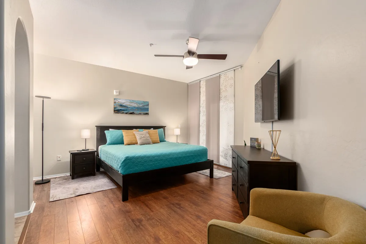 This interior image showcases a serene bedroom setting with a bed dressed in a turquoise cover, ocean-themed artwork above the headboard, stylish nightstands with lamps, a modern wall-mounted TV, and a cozy reading chair. The tranquil and cozy mood aligns with a restful master suite atmosphere.
