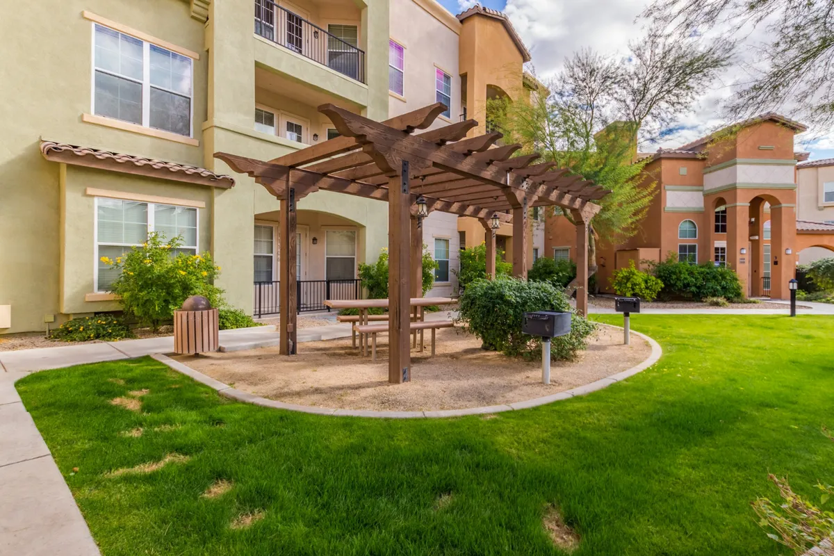 A charming pergola shades a picnic table set amid green grass and surrounded by community buildings. The area invites residents to gather for picnics, celebrate special occasions, or simply enjoy quiet moments outdoors.