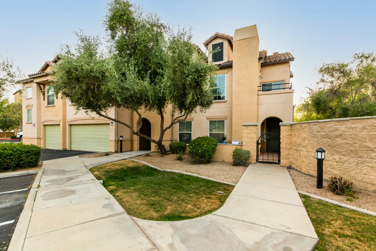 This image shows the exterior of the condo, featuring the inviting entrance pathway shaded by a mature olive tree, the building’s facade, and garage doors. The landscaped front area is tranquil and welcoming, perfect for morning walks or sipping coffee outdoors.
