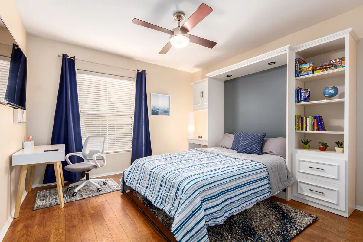 An inviting, light-filled bedroom doubles as a functional workspace, featuring a bed with striped bedding, built-in bookshelves, a desk with chair, and a window streaming in daylight. The room feels both calm and practical, ideal for remote work or relaxation.