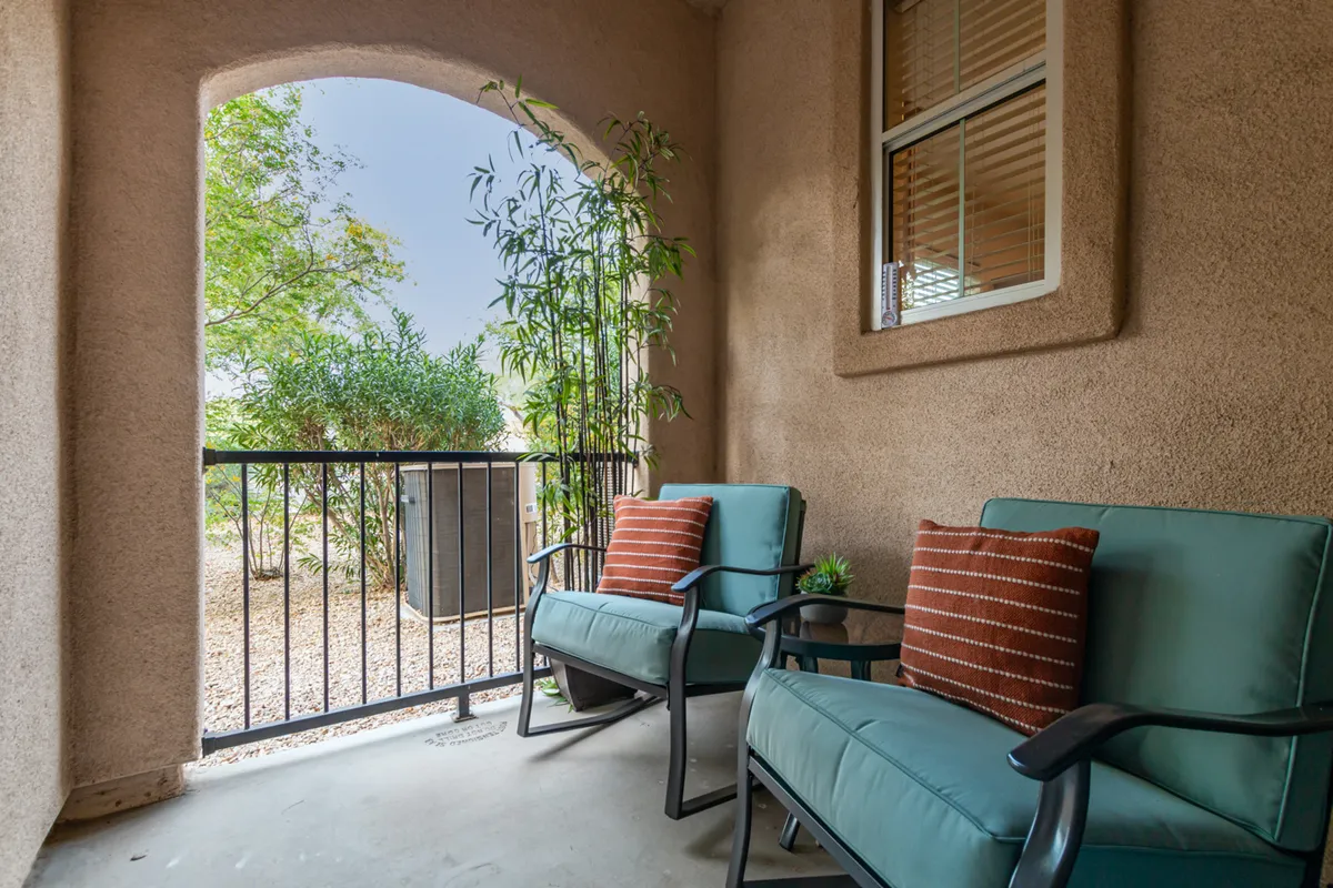 A cozy balcony setup with two comfortable chairs, decorative pillows, potted plants, and a railing overlooking the community. The tranquil mood suggests morning coffee or evening conversations, with a blend of privacy and connection to the outdoors.