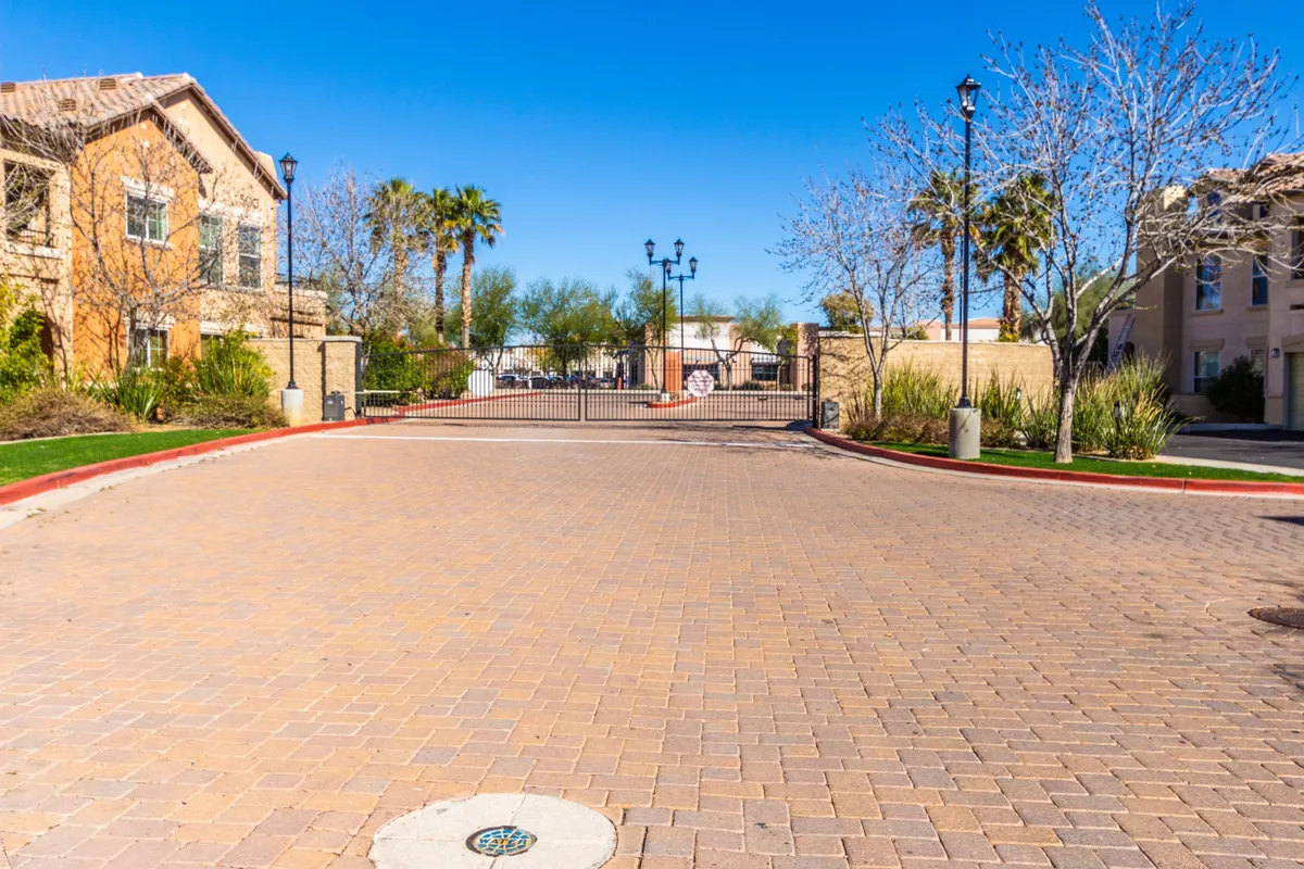 The gated entrance to Park Place is showcased here, with a paved pathway, mature landscaping, and street lamps lining the way. Neighbors are visible enjoying a morning walk and chatting, creating a sense of community and security.
