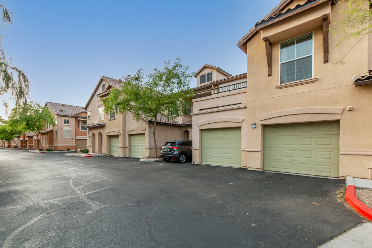 This image displays the townhouse architecture with green garage doors, surrounded by well-maintained landscaping. The scene captures a resident strolling along the path and hints of social interaction in the common outdoor area.